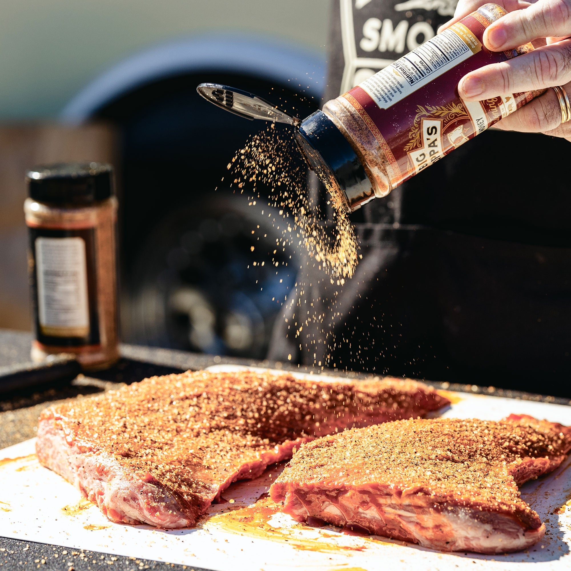 Big Poppa sprinkling Double Secret Steak Seasoning on two tri-tips pieces of meat that are generously coated.