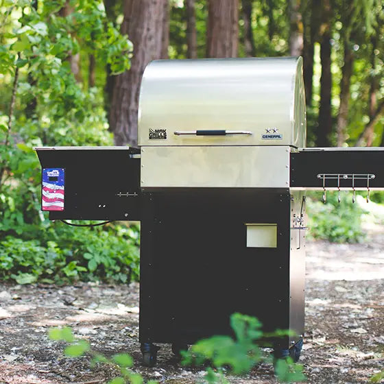 MAK Grills General smoker with stainless lid, black base, and U.S. flag decal, set up outdoors in a forested area.