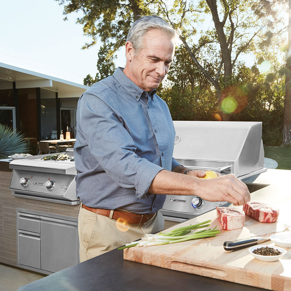 An older man in a blue shirt prepares food outdoors next to a stainless steel grill. He is seasoning two raw steaks on a wooden cutting board with green onions and peppercorns. A modern outdoor kitchen and a sunlit garden are visible in the background.