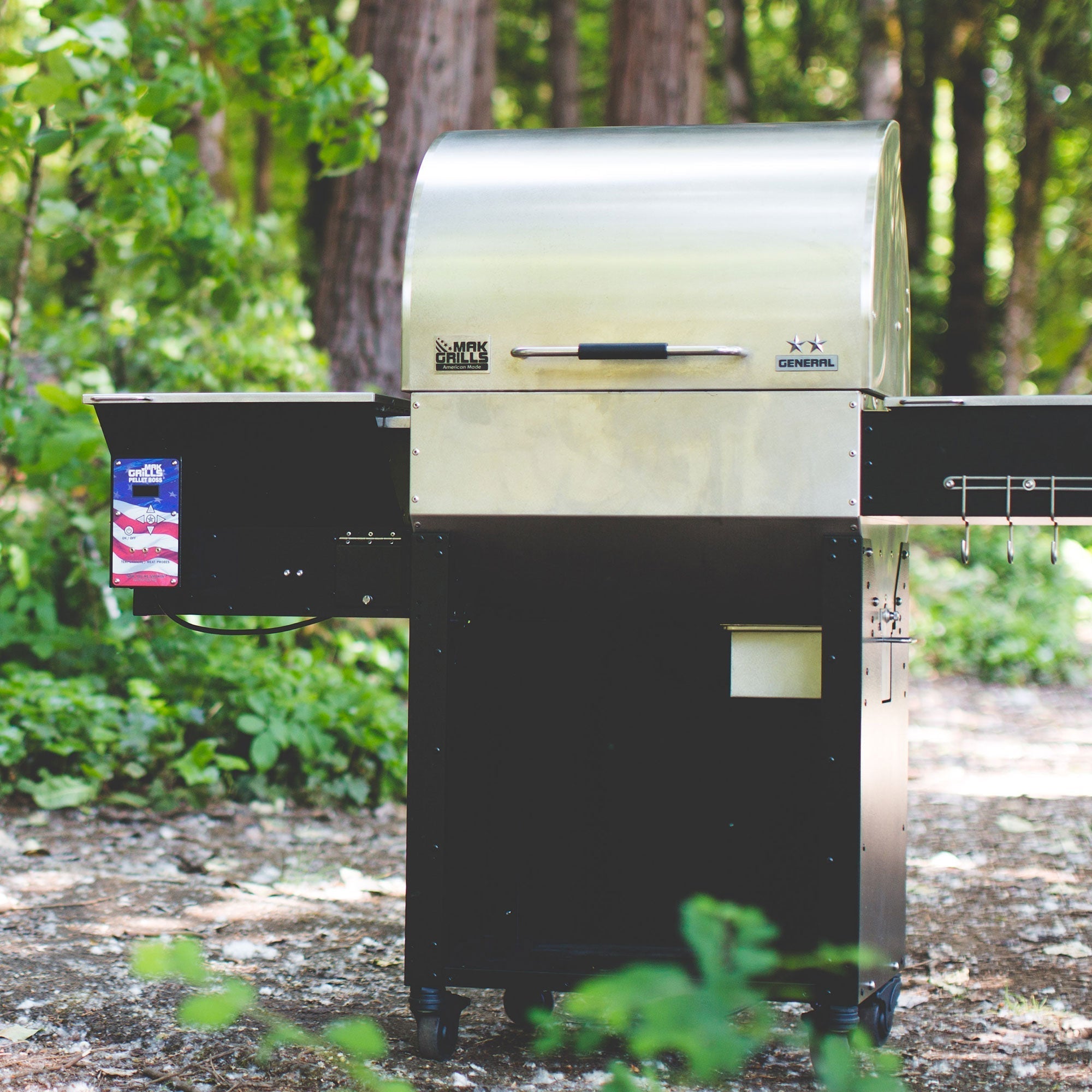 A MAK Grills barbecue smoker, model "General," set up in an outdoor forested area. The smoker has a stainless steel lid and black base, with the American flag decal on the side hopper.