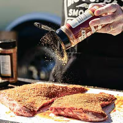 Big Poppa sprinkling Double Secret Steak Seasoning on two tri-tips pieces of meat that are generously coated.