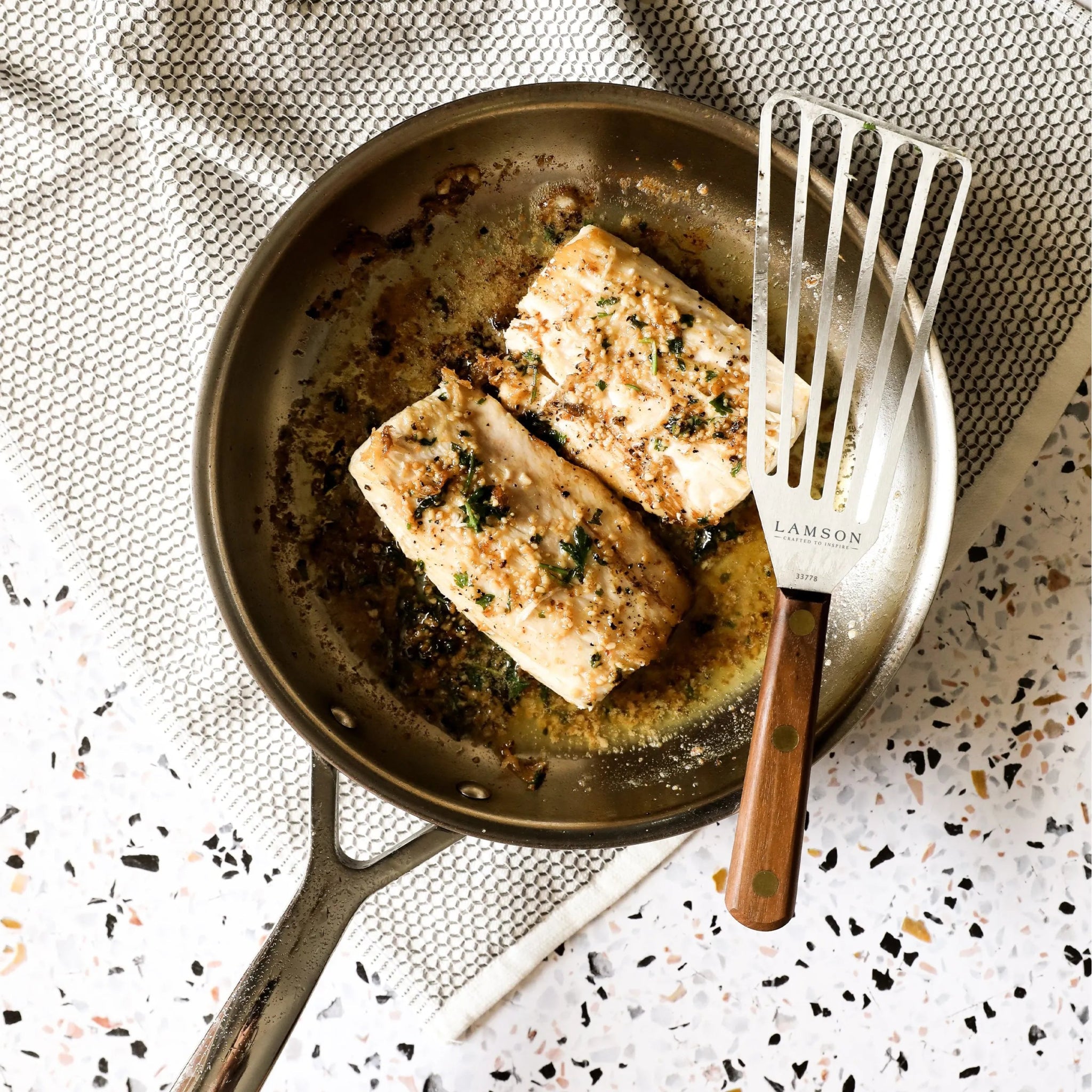 Sauce pan that features two pieces of salmon and the perforated turner to the side to be used to flip the fish.