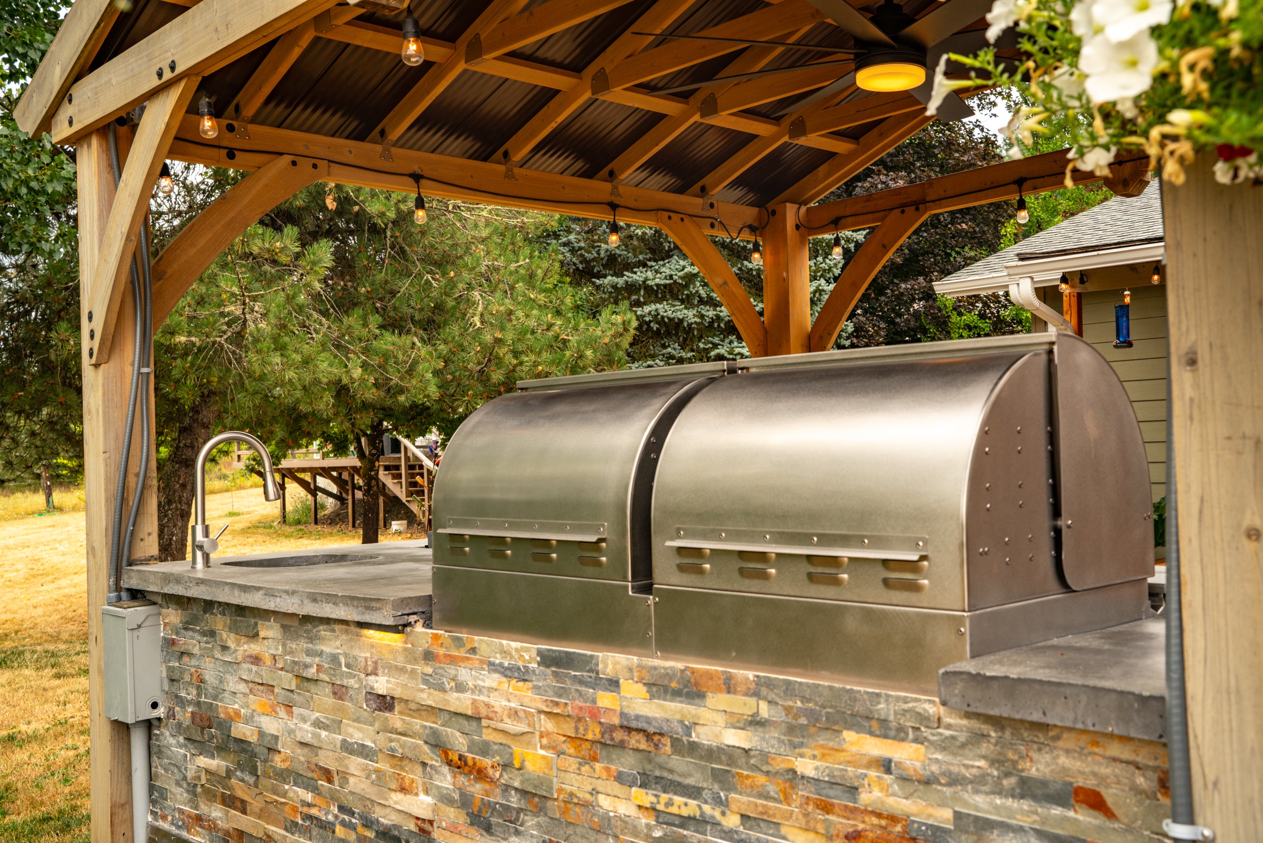 Back view of a MAK Grills pellet smoker and built-in grill in an outdoor kitchen, under a wooden pergola, with a faucet and surrounding greenery.
