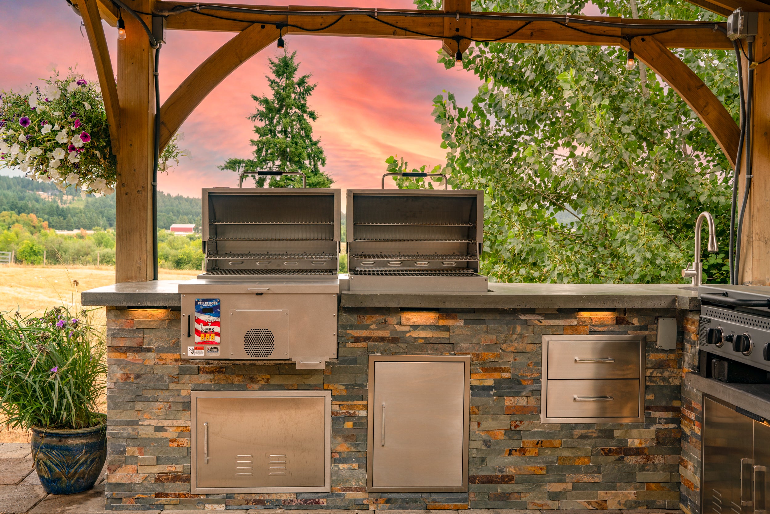 Outdoor kitchen with an open MAK Grills pellet smoker and grill, showcasing the grilling area with stainless steel grates, set against a backdrop of trees.