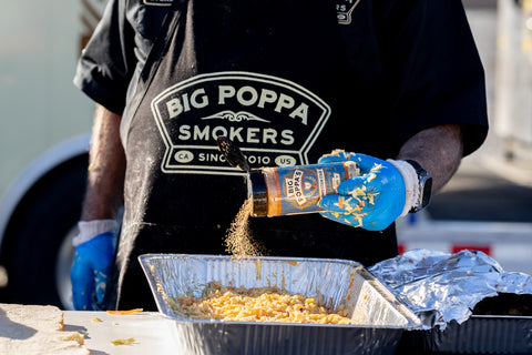 A man in a black apron labeled 'Big Poppa Smokers' seasoning a dish with Big Poppa's spice blend from a bottle, during a competitive cooking event. He wears blue gloves and the background shows an outdoor cooking setting.