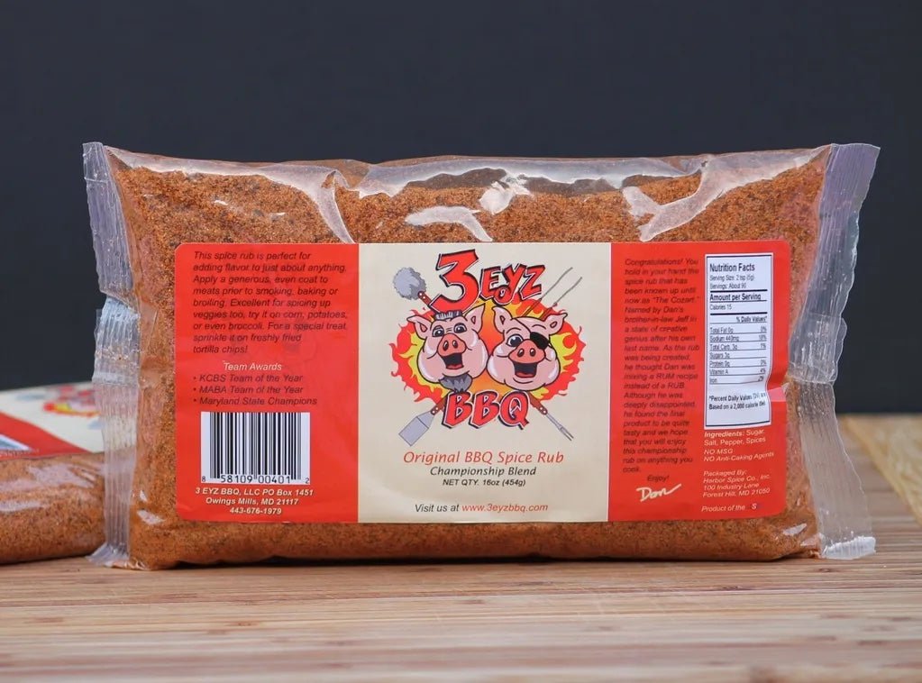 3 Eyz BBQ Original BBQ Spice Rub in a clear plastic bag with a label featuring three cartoon pigs, placed on a wooden surface against a plain background.