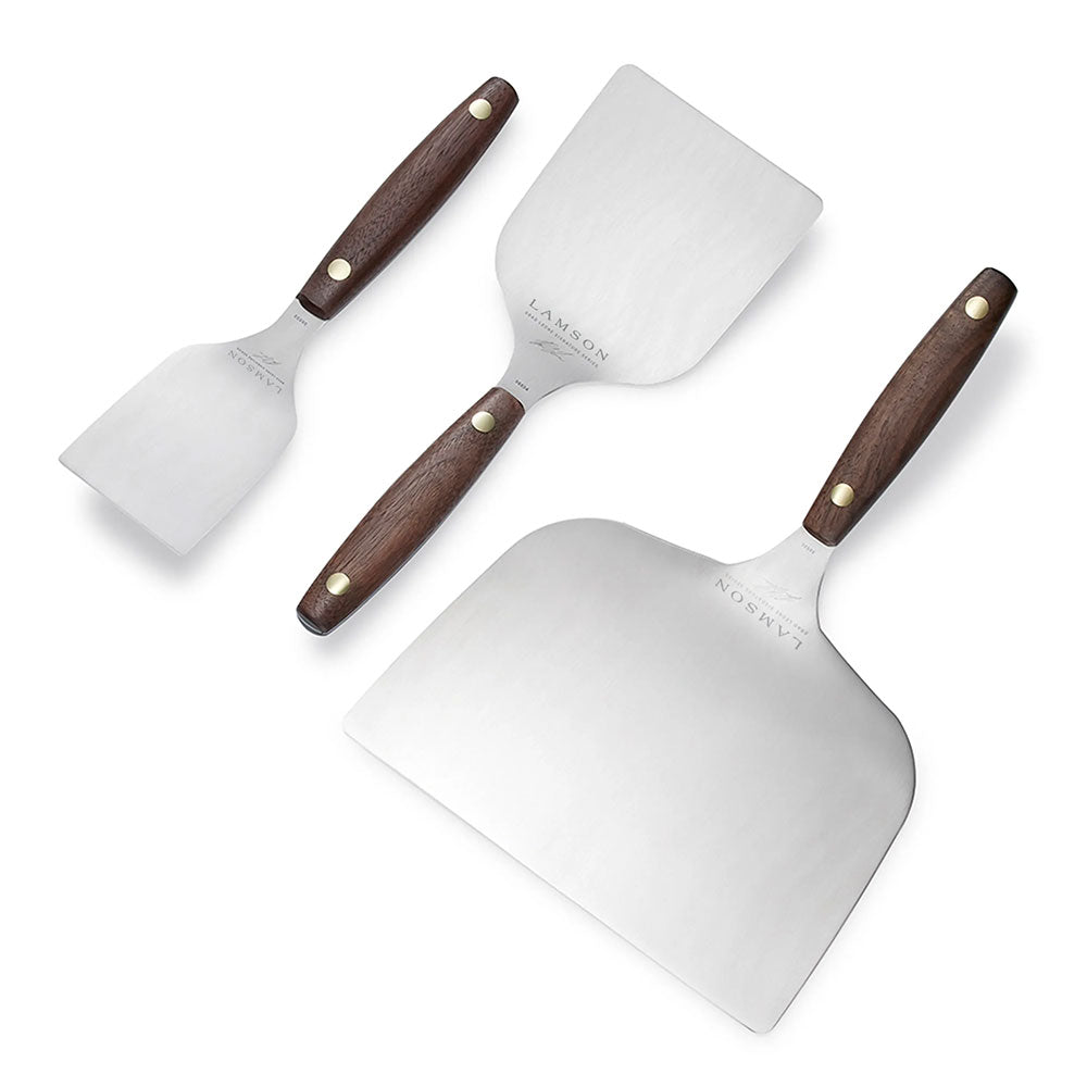 A set of three Lamson griddle turners with stainless steel blades and wooden handles featuring brass rivets. The turners are laid out on a white background, with the largest turner at the bottom right, the medium-sized turner at the top right, and the smallest turner at the left. Each turner has a sleek, polished finish and ergonomic design.