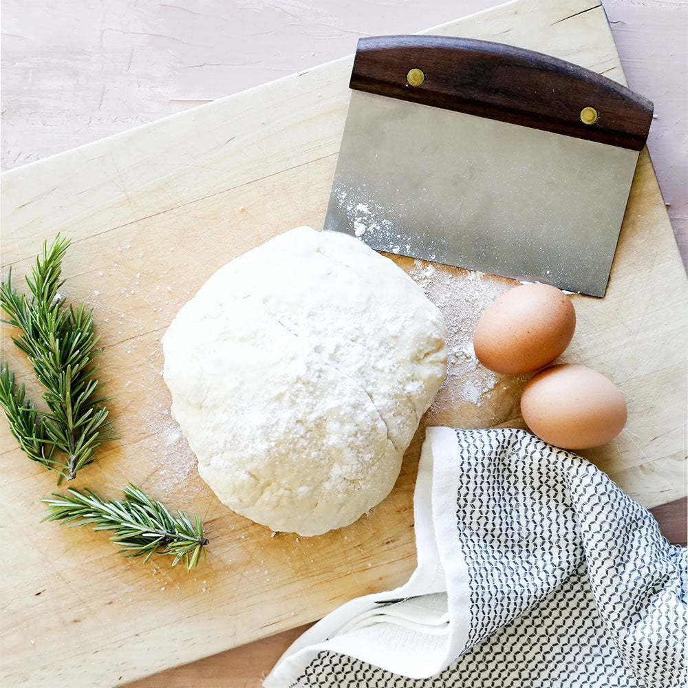 A wooden cutting board with a ball of dough dusted with flour, two brown eggs, a sprig of rosemary, and a metal dough scraper with a wooden handle. A white and black checkered cloth is partially visible at the bottom right.