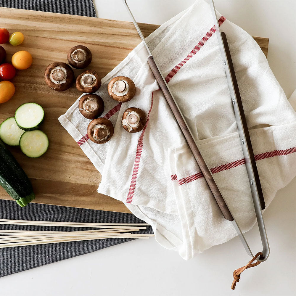 A wooden cutting board with a white and red striped cloth, six brown mushrooms, and a pair of long-handled tongs. To the left of the cutting board are sliced zucchini, cherry tomatoes, and wooden skewers, placed on a dark placemat.