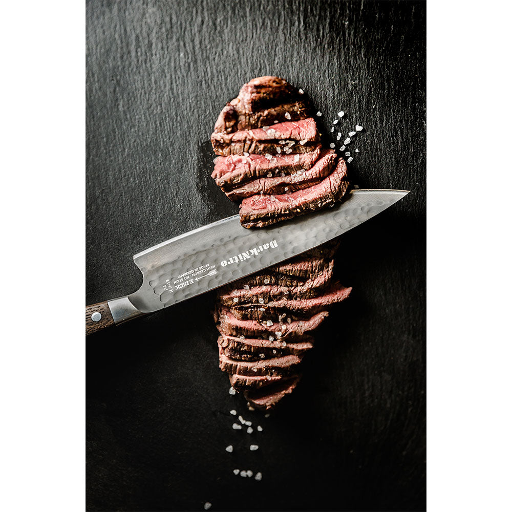 A DarkNitro chef's knife with a hammered blade finish slices through a piece of grilled steak, displayed on a black slate surface. Coarse salt is sprinkled around the steak.