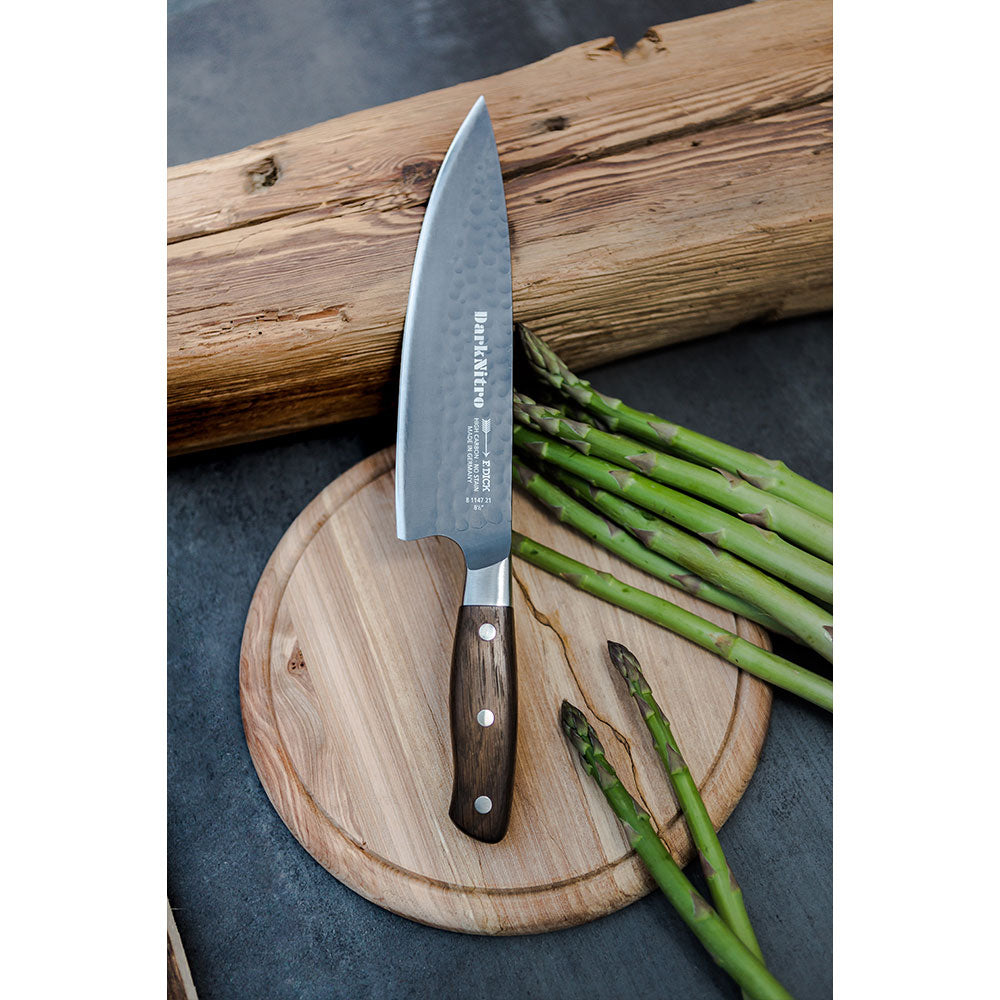 A DarkNitro chef's knife with a hammered blade finish rests on a round wooden cutting board alongside a bundle of asparagus spears. The background features a rustic wooden log.