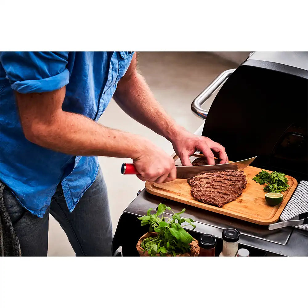 Man slicing grilled steak on wooden cutting board with red-handled knife next to herbs and a lime wedge.