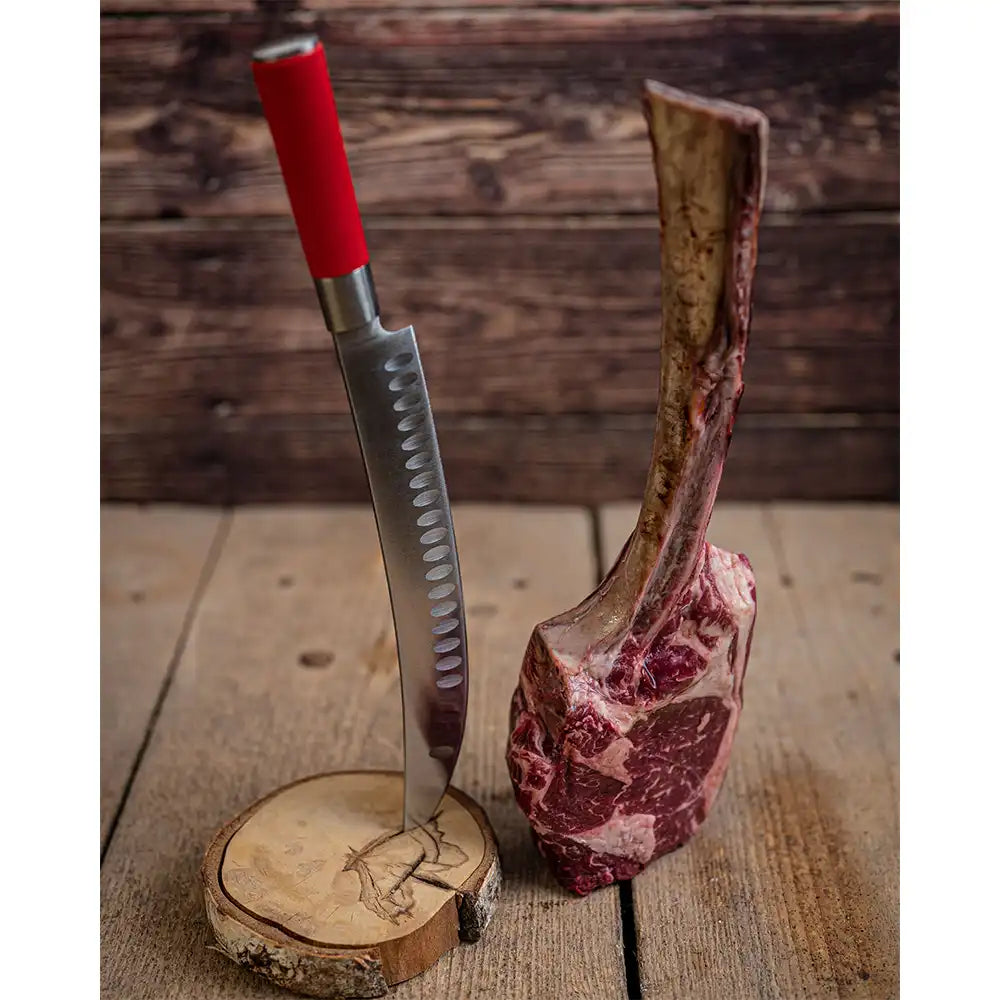 Raw tomahawk steak standing upright beside a red-handled knife stuck into a wood round.