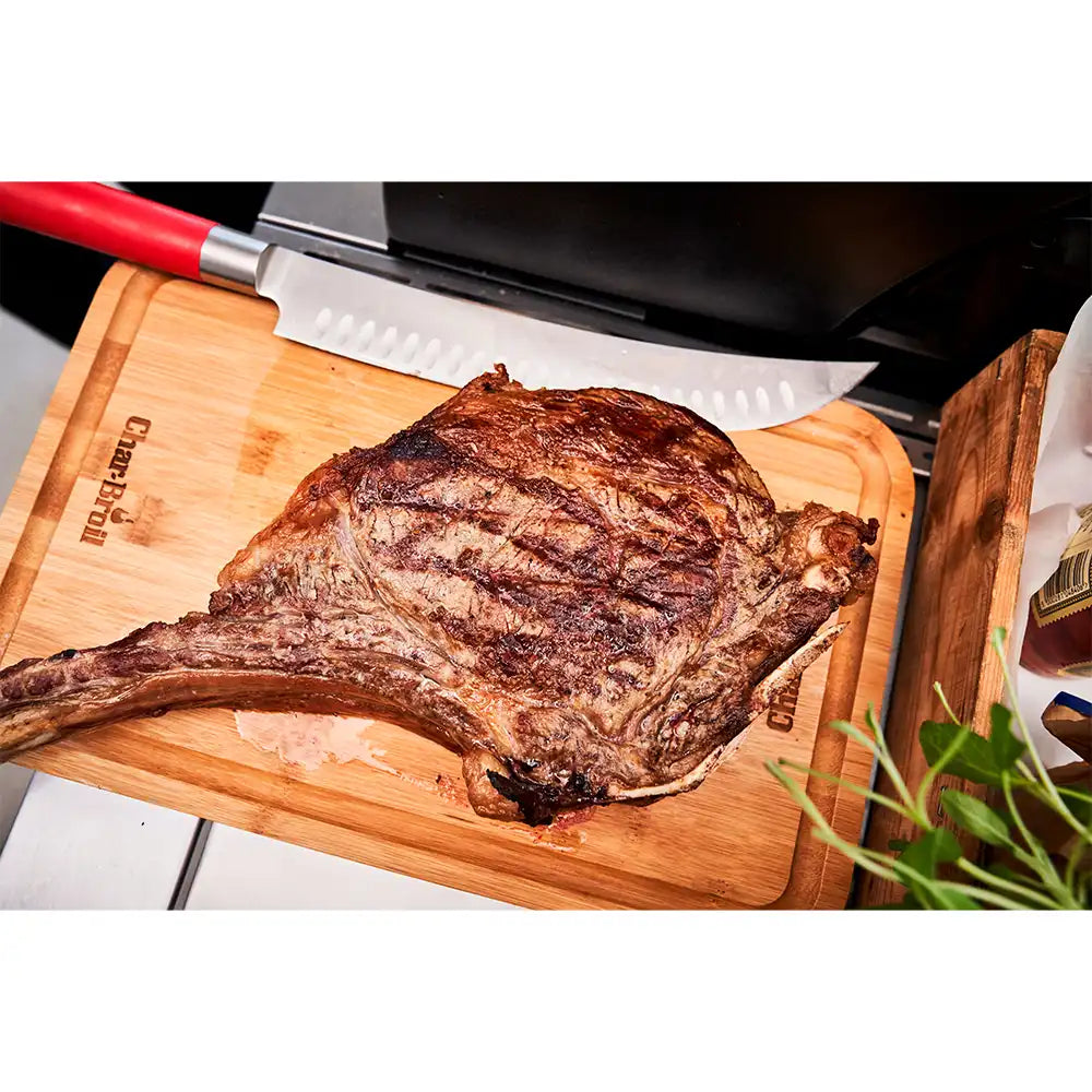 Grilled tomahawk steak resting on a Char-Broil cutting board next to a red-handled carving knife.
