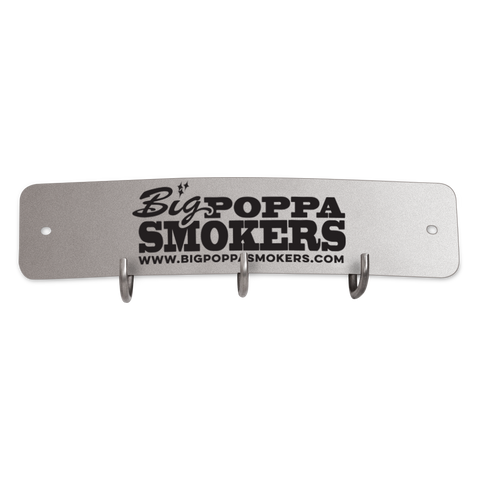 A rectangular metal tool hook shield featuring the logo of Big Poppa Smokers. The shield has a silver-grey finish with the logo in black, including the website 'www.bigpoppasmokers.com' printed below. It includes three hooks for hanging tools, centrally located along the bottom edge.