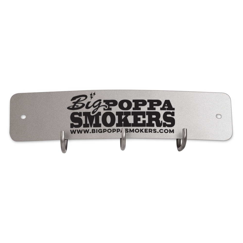 A rectangular metal tool hook shield featuring the logo of Big Poppa Smokers. The shield has a silver-grey finish with the logo in black, including the website 'www.bigpoppasmokers.com' printed below. It includes three hooks for hanging tools, centrally located along the bottom edge.