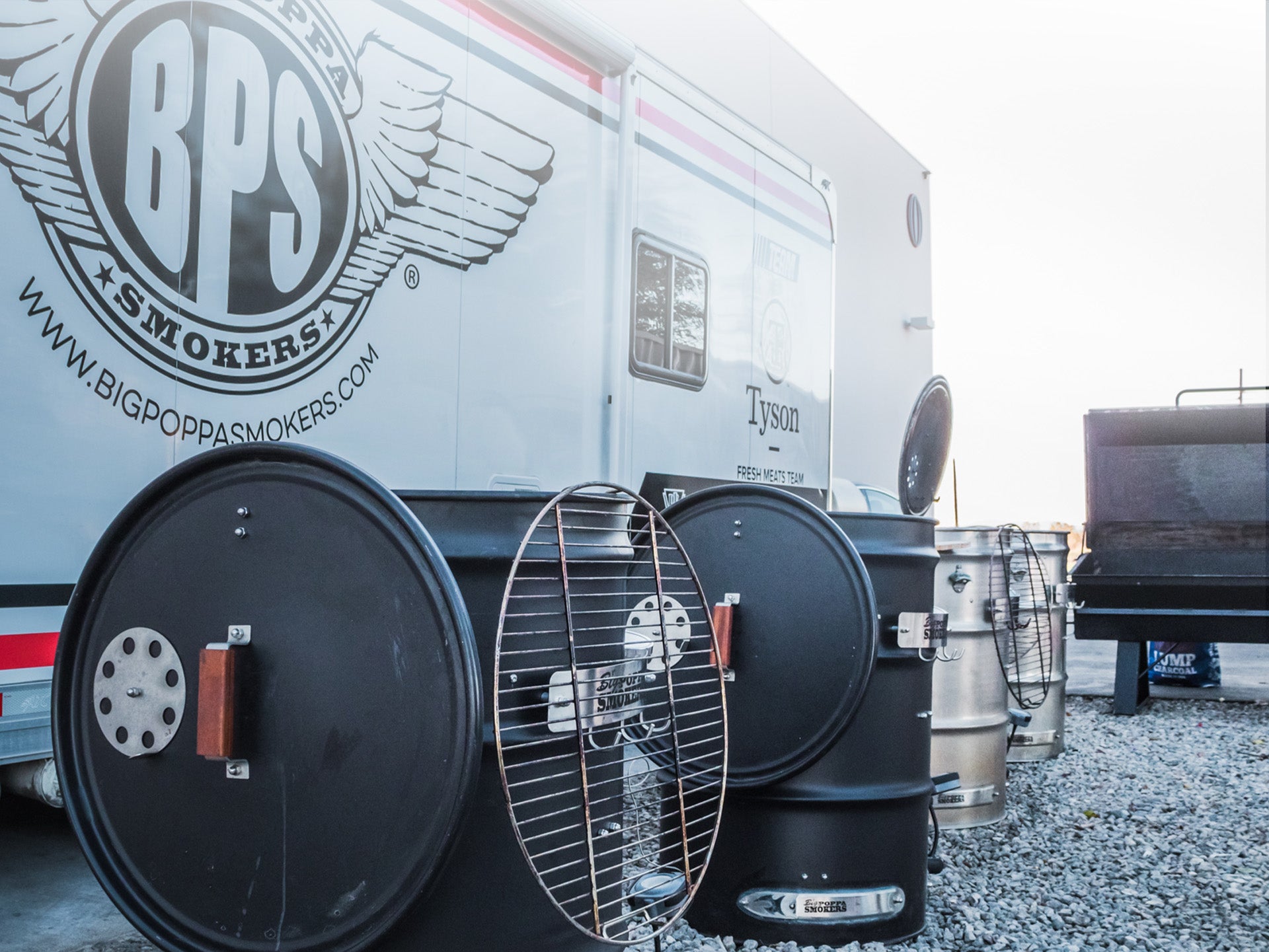 A row of black metal barrel smokers with wooden handles are lined up outside in front of a large white truck with "Big Poppa Smokers" branding.