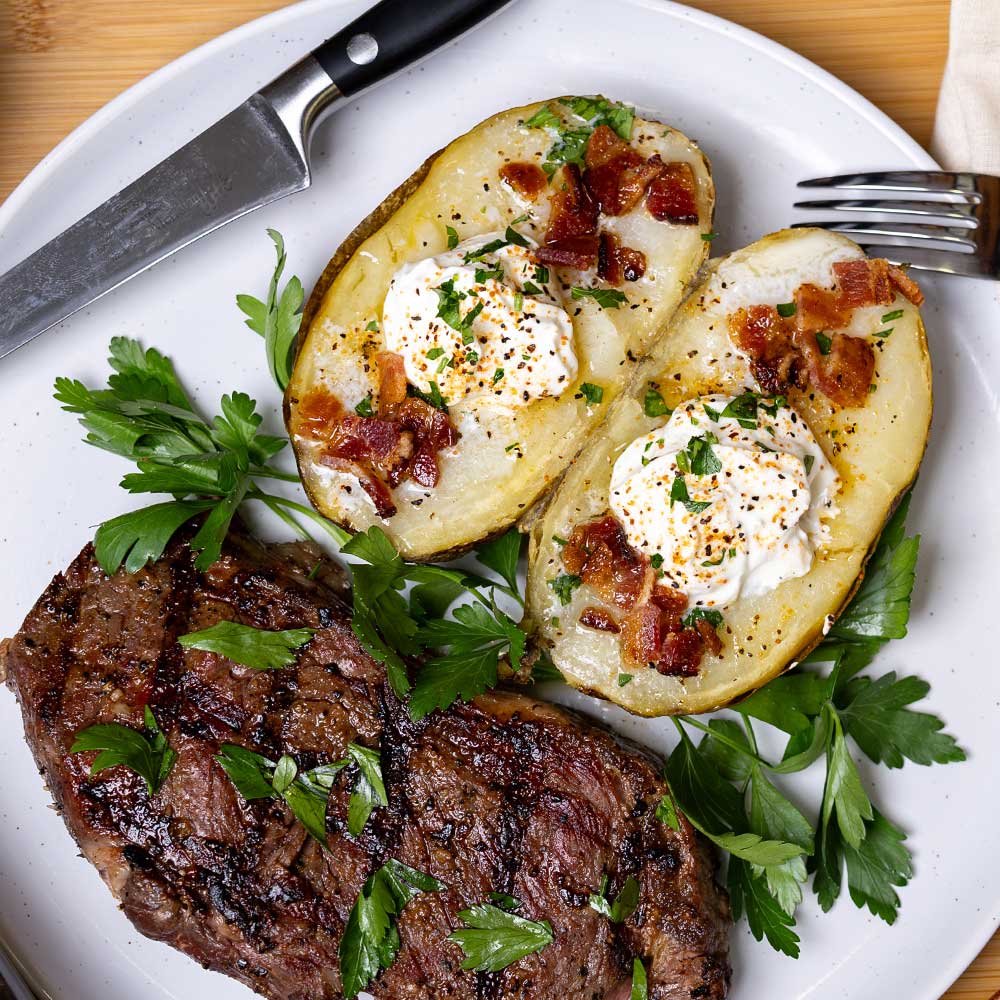 steak with perfect grill marks plated next to buttered grilled potatoes topped with chopped bacon and sour cream, garnished with a pop of green parsley on a white plate with a fork and knife resting on the white plate.