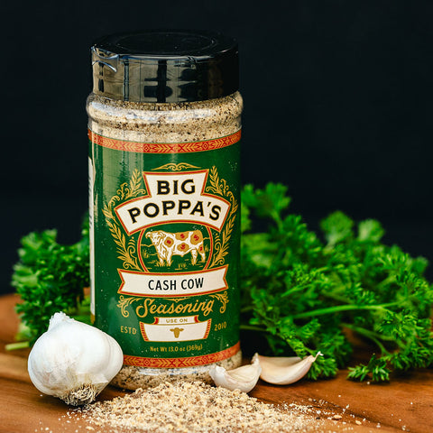 A bottle of 'Big Poppa's Cash Cow' seasoning placed on a dark wooden surface, accompanied by fresh parsley and garlic cloves. The background is dark, emphasizing the green bottle label decorated with cows and elegant patterns.