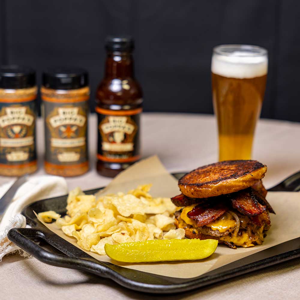 mouthwatering display of a smash burger on a skillet with chips and a pickle, the burger of which is seasoned to perfection with competition brisket and steak seasoning and stacked high with thick cut bacon, grilled onions, and cheese