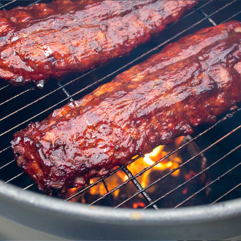 juicy ribs drenched in barbecue sauce shown being grilled in a drum smoker over flaming charcoals