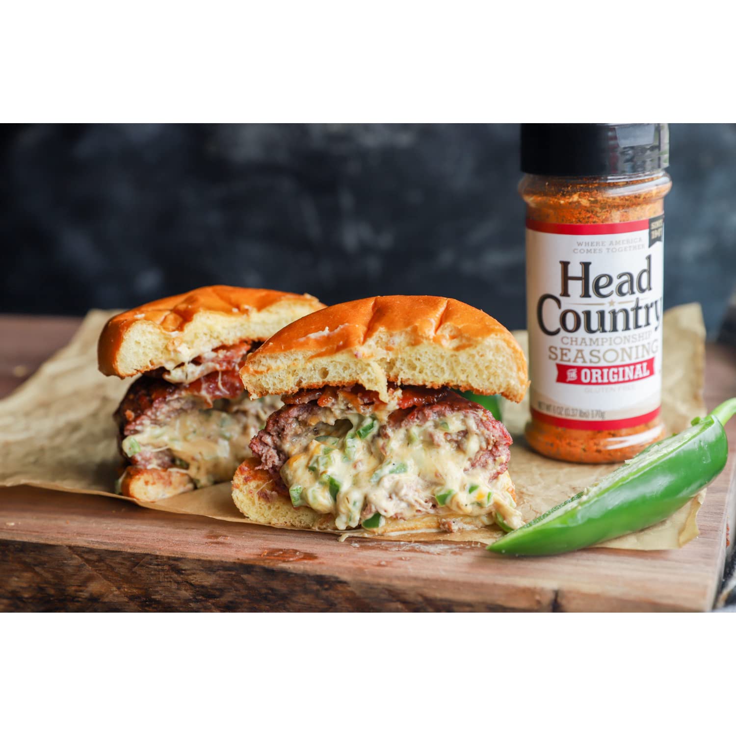 A juicy burger cut in half, filled with melted cheese and jalapeños, is placed on a wooden surface next to a bottle of Head Country Championship Seasoning. A green chili pepper is also visible beside the burger.