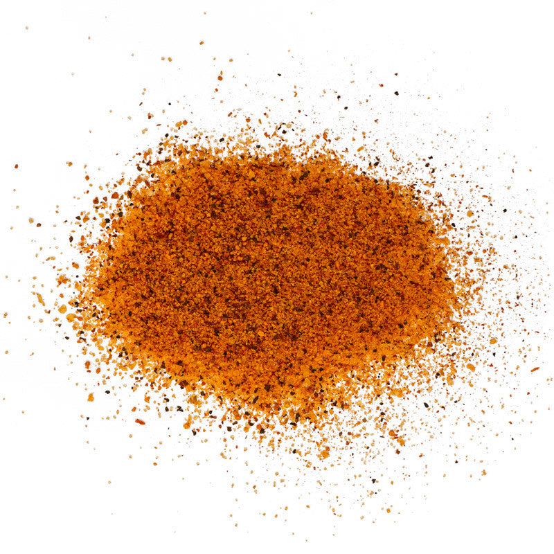 A close-up view of the JDQ BBQ Rub powder spread out on a white surface. The seasoning is a coarse mixture, predominantly orange with visible black pepper flakes and other spices.