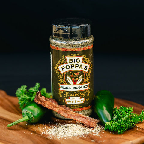 A bottle of 'Big Poppa's Jallelujah Jalapeño Bacon Seasoning' next to a fresh jalapeño and a strip of cooked bacon, with parsley and seasoning dust scattered on a wooden board, emphasizing the seasoning’s flavor profile.