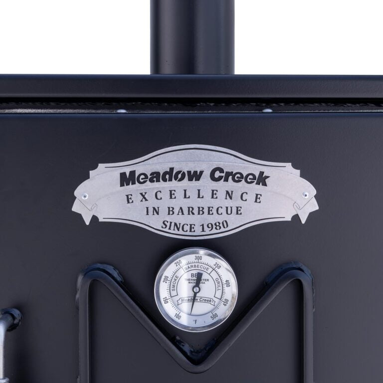 Close-up of the thermometer and logo on the upper door of the Meadow Creek BX25 smoker, displaying the text 'Meadow Creek Excellence in Barbecue Since 1980' above the temperature gauge.