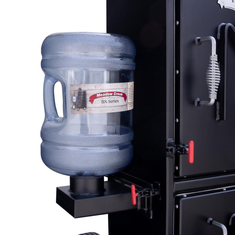 Close-up of the water bottle attached to the Meadow Creek BX25 smoker, featuring a large transparent bottle labeled 'Meadow Creek BX Series.