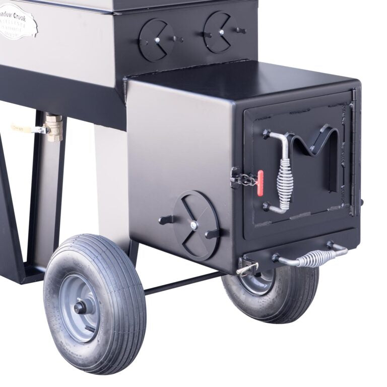 Side view of a Meadow Creek smoker with the firebox door closed. The smoker is equipped with large wheels, air vents, and a spring handle for safe operation. The manufacturer's logo is displayed on the main cooking chamber.