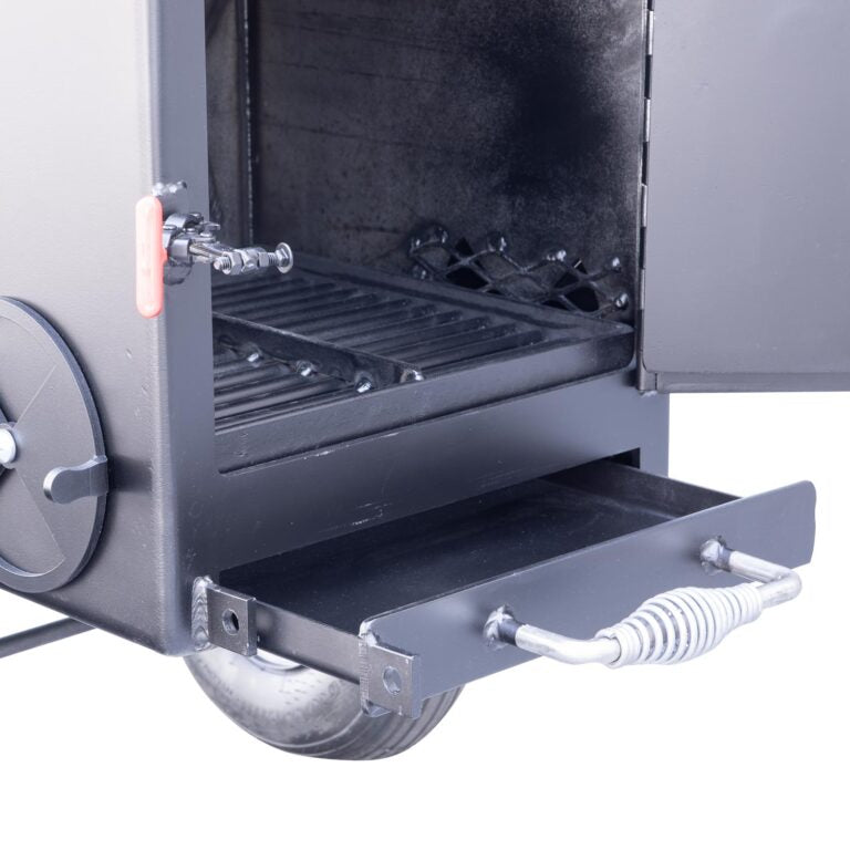 Close-up view of the open firebox of a Meadow Creek smoker, showing the ash pan partially pulled out. The interior is blackened from use, with a red handle on the left side for adjusting the airflow.