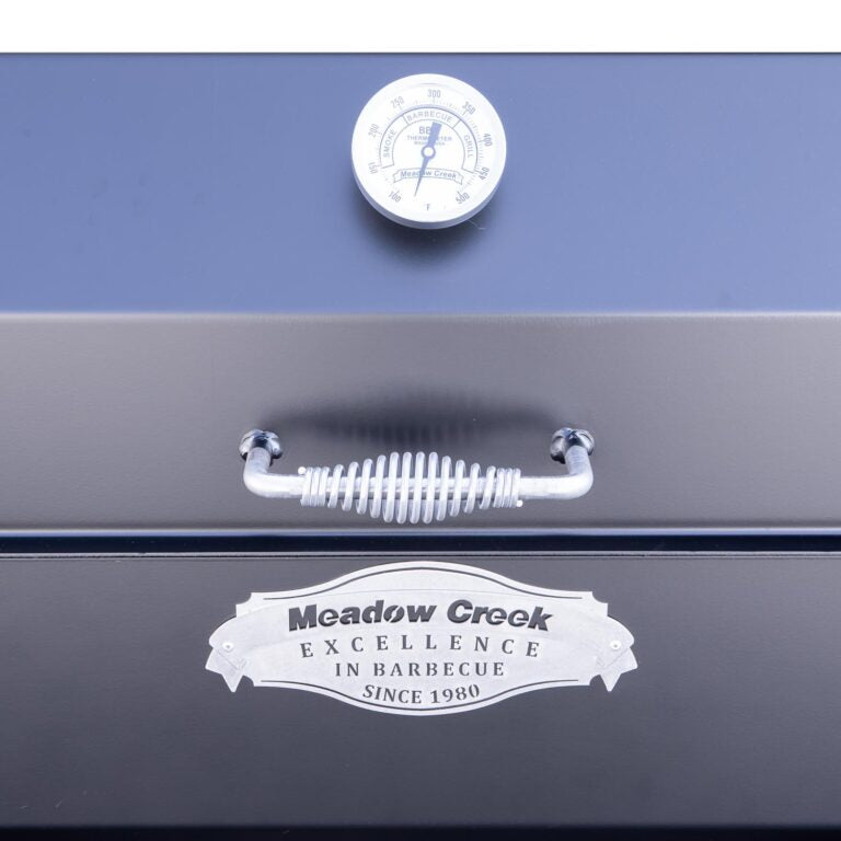 Close-up of the thermometer and spring handle on the lid of a Meadow Creek smoker. The thermometer shows the temperature in both Fahrenheit and Celsius, and the logo below reads 'Meadow Creek, Excellence in Barbecue, Since 1980.