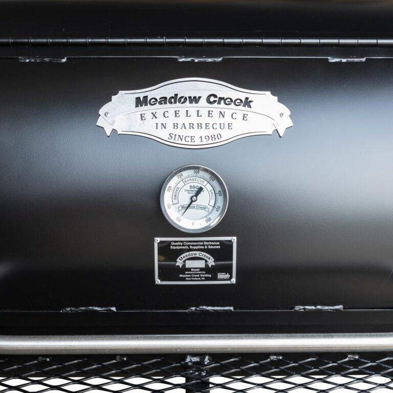 Close-up view of the front of a Meadow Creek barbecue smoker. The black metal surface displays a silver badge reading "Meadow Creek Excellence in Barbecue Since 1980." Below the badge, there is a circular thermometer showing temperature measurements. An additional plaque indicates the model and manufacturing details.