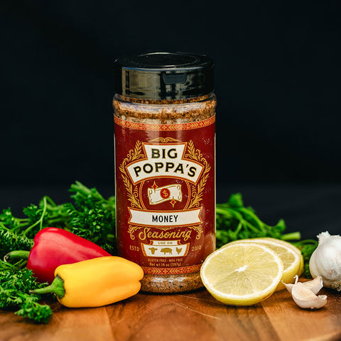 A bottle of 'Big Poppa's Money Seasoning' displayed with vibrant red and yellow bell peppers, garlic, lemon slices, and fresh parsley on a dark wooden surface, highlighting its use in cooking.