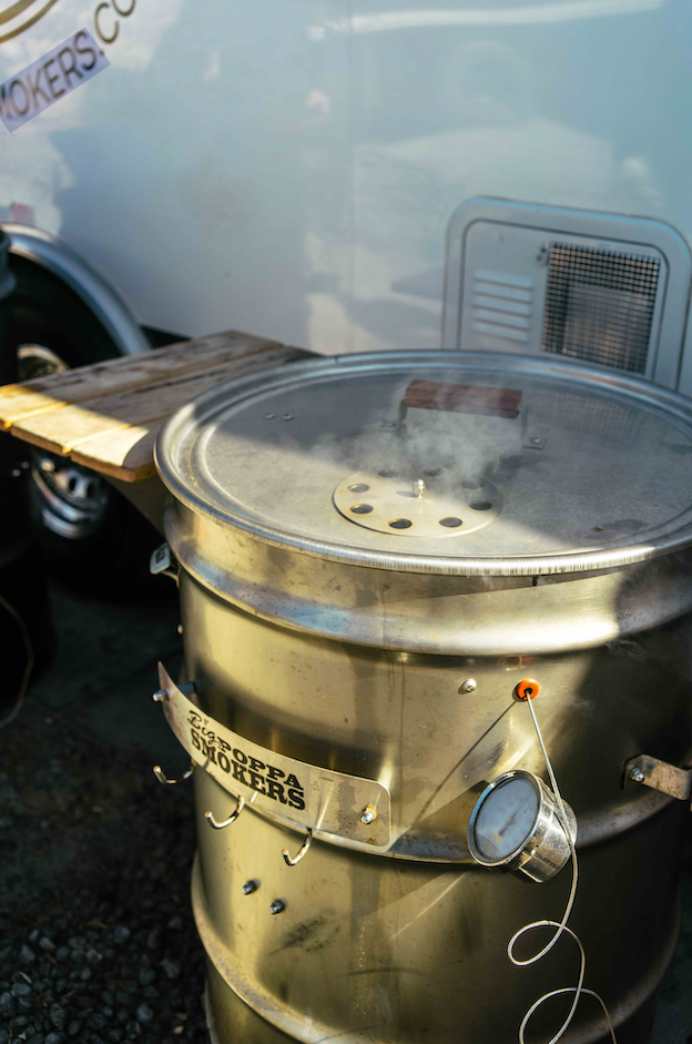 A metal smoker with a wooden handle on its lid, emitting smoke. The smoker has the "Big Poppa Smokers" logo on the front and a temperature gauge attached to the side. The background includes a white trailer with some text partially visible.