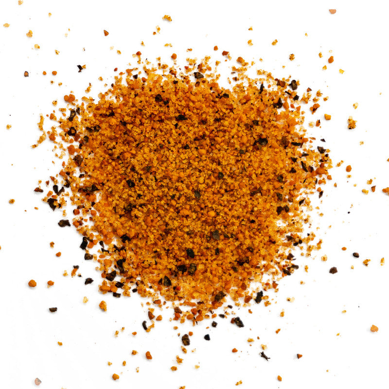 A close-up image of Simply Marvelous Season All rub. The seasoning consists of a mix of orange and black coarse spices spread out on a white surface.