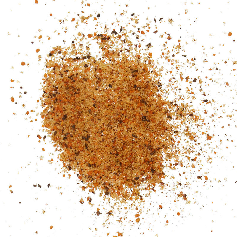 A detailed close-up of Smokin' Guns BBQ Hot Rub, showing a mix of fine granules in various shades of brown, orange, and black, scattered across a white surface.