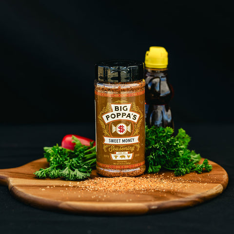A bottle of Big Poppa's Sweet Money Seasoning on a wooden cutting board with fresh herbs and condiments.