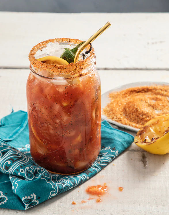 A mason jar filled with a mixed beverage containing ice, lemon slices, and a spicy rim, garnished with a lemon wedge and a jalapeño slice. The jar is placed on a blue patterned cloth, with a plate of seasoning and a lemon half in the background.