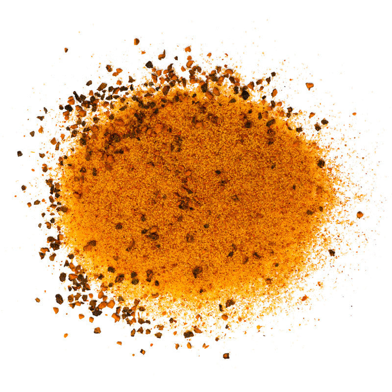 Close-up view of Williams Rib Tickler BBQ Rub. The seasoning is a mix of fine orange-brown powder with larger dark brown flakes, scattered on a white surface.