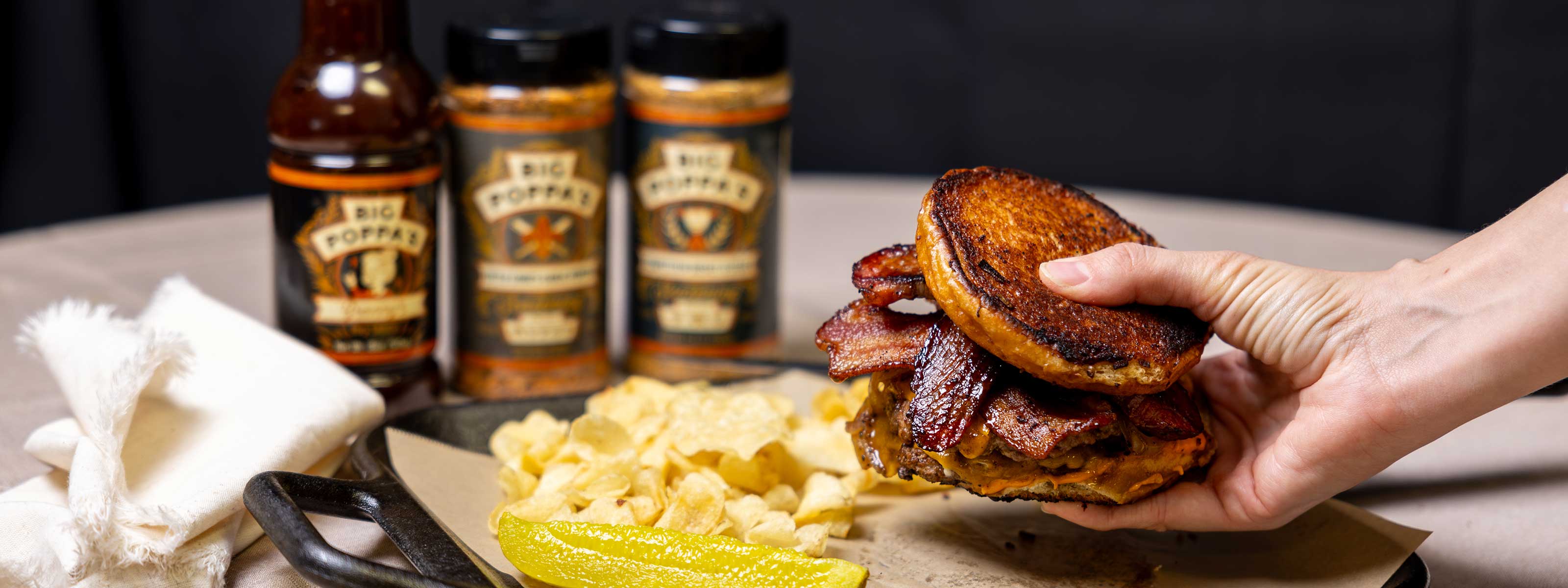 A hand holding a bacon cheeseburger with BBQ Sauce and Seasonings in back