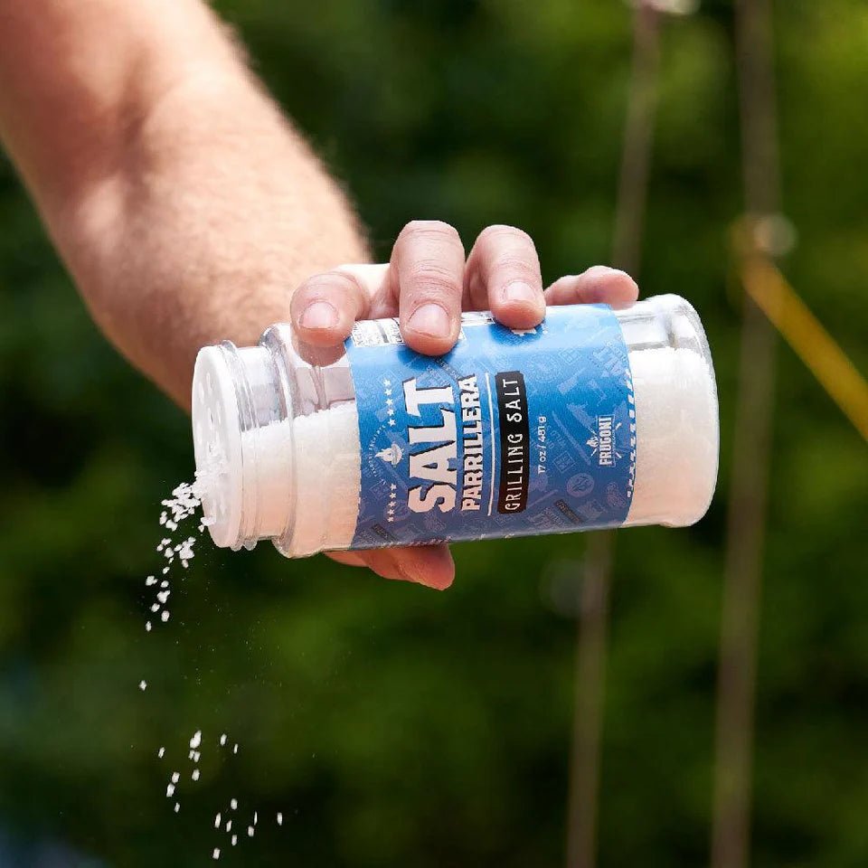 A person is shaking Frugoni Salt Parrillera grilling salt out of a jar. The jar is tilted, and salt is visibly falling out. The background is green, suggesting an outdoor setting.