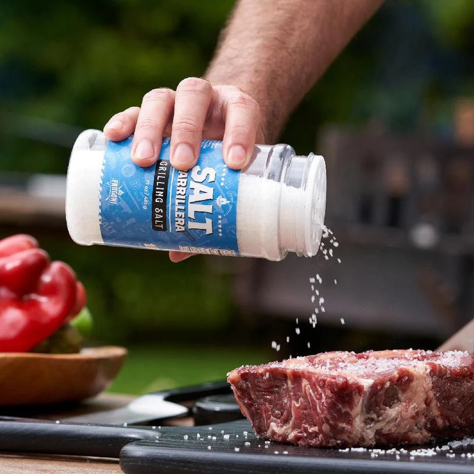 A person is pouring Frugoni Salt Parrillera grilling salt onto a raw steak on a black cutting board. A red bell pepper is visible in the background on the left, and the scene is set outdoors.