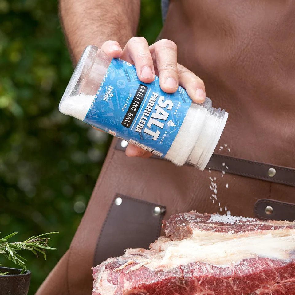 A person is sprinkling Frugoni Salt Parrillera grilling salt onto a thick cut of raw meat. The person is wearing a brown apron, and the scene appears to be set outdoors with greenery in the background.