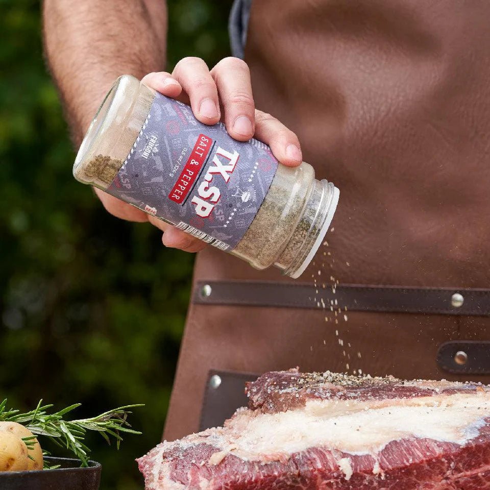 A close-up of a hand seasoning a large piece of meat with 'TX.SP Salt & Pepper' from a container. The person is wearing a brown leather apron, and there are herbs and potatoes in the background.