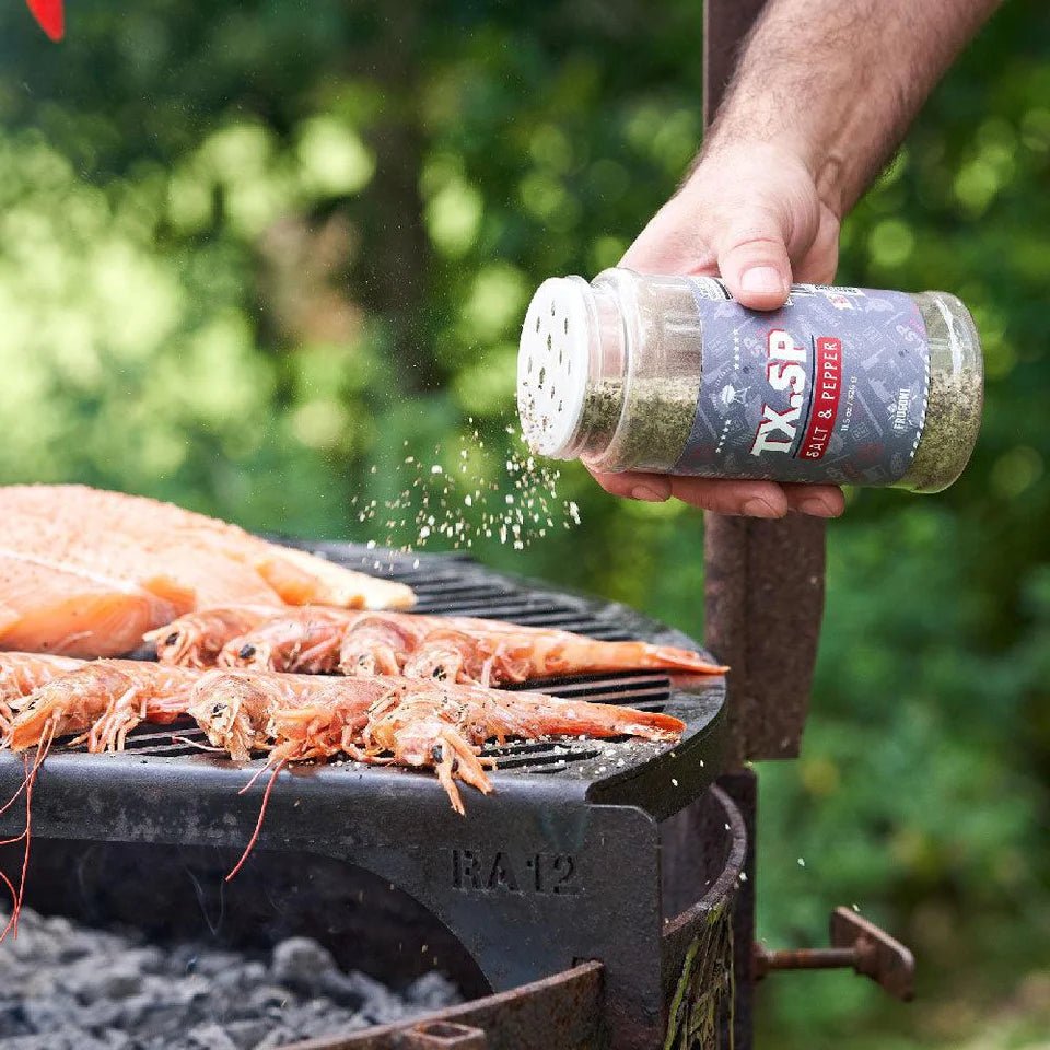 A hand sprinkling 'TX.SP Salt & Pepper' seasoning from a container over shrimp and salmon cooking on an outdoor grill. The background is blurred with greenery.