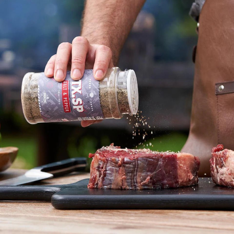 A close-up of a hand sprinkling salt and pepper seasoning from a large container labeled 'TX.SP Salt & Pepper' onto a thick cut of raw steak on a wooden cutting board. A knife is visible in the background.