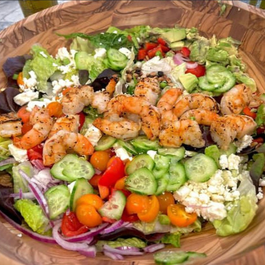 A colorful salad featuring grilled shrimp on top, with mixed greens, cherry tomatoes, cucumbers, red onions, and crumbled cheese in a large wooden bowl.