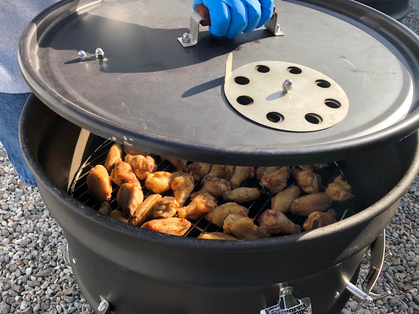 A person using blue-gloved hands to handle chicken pieces in a black drum-style outdoor grill filled with various meats, showing an open cooking setup.